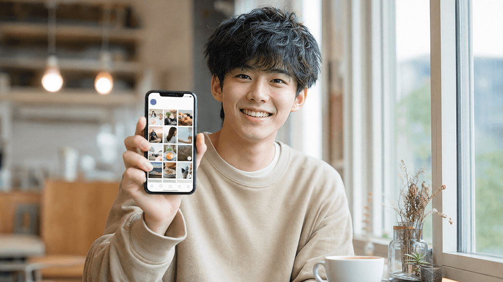 カフェの窓辺でスマートフォンにSNSのフィードを表示し笑顔の日本人。REDアプリを始めてみるイメージ